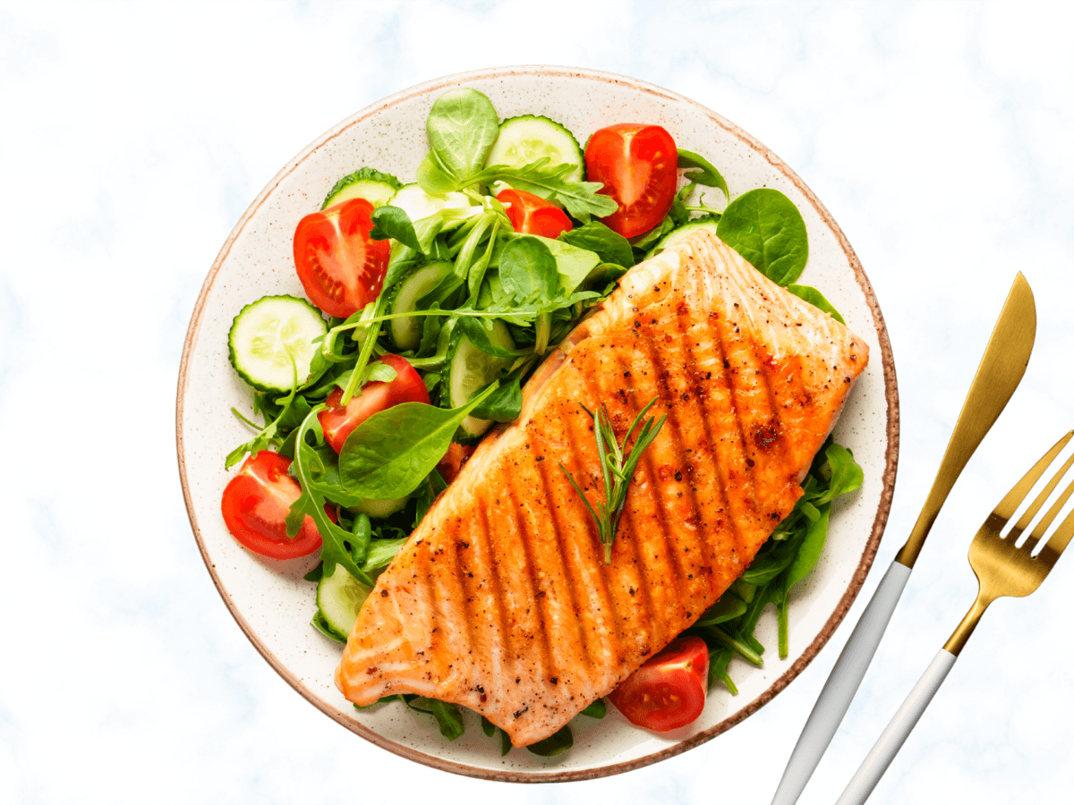 Grilled salmon fillet and fresh salad with cucumber and tomatoes on a plate, with a fork and knife.